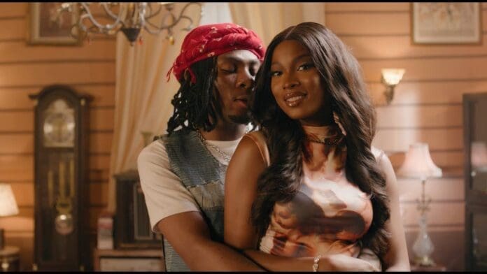 A man stands behind a smiling woman, embracing her in a warmly lit room with vintage decor and soft lighting, as Joeboy's melodies fill the air.