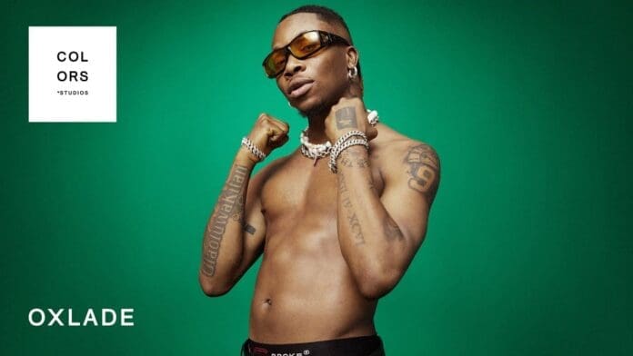 A man with tattoos on his arms poses confidently, capturing the vibrant energy of COLORS SHOW and the rhythm of Oxlade's hit "KU LO SA.