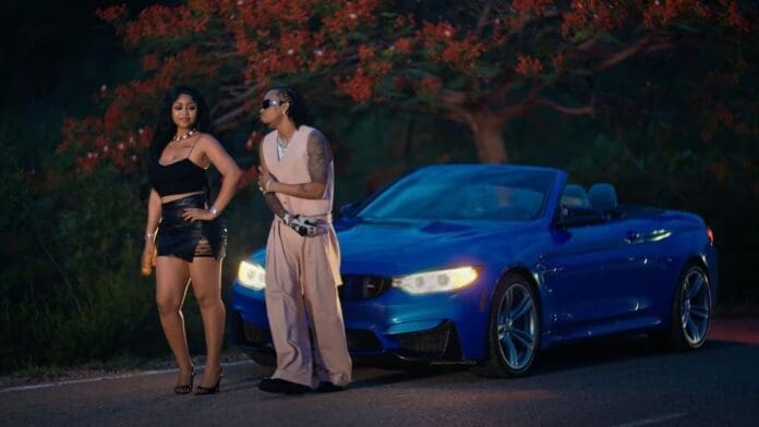 A woman in a black outfit and a man in light-colored clothes stand beside a blue convertible car, reminiscent of a Tekno Official Video, parked on a road with trees in the background.