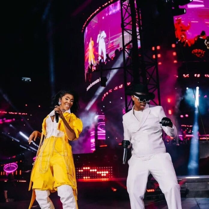 Two performers from Mafikizolo sing and dance on stage under bright lights, celebrating South African heritage, as large digital screens display their images in the background.