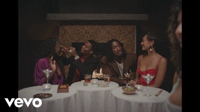 Four people sit at two round tables with drinks, two interacting closely, while another pair sits side by side; a cozy, dimly lit setting reminiscent of Nights In The Sun, with a fireplace glowing in the background.