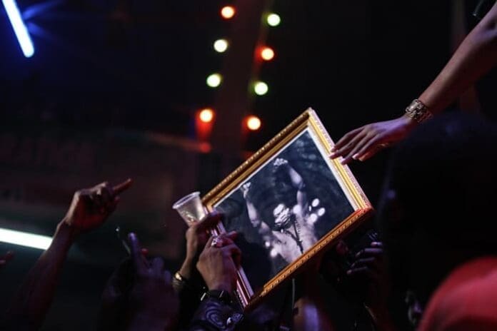 A crowd of people raise a framed black-and-white photo of Fela Kuti, with hands reaching toward it and colorful lights in the background, celebrating his impact on African music and his Grammy lifetime achievement award.