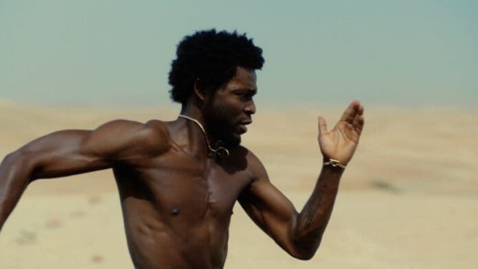 A shirtless man runs outdoors in a desert landscape under clear skies, his right arm raised and his face focused ahead, capturing the vibrant energy of an Official Music Video by Omah Lay.