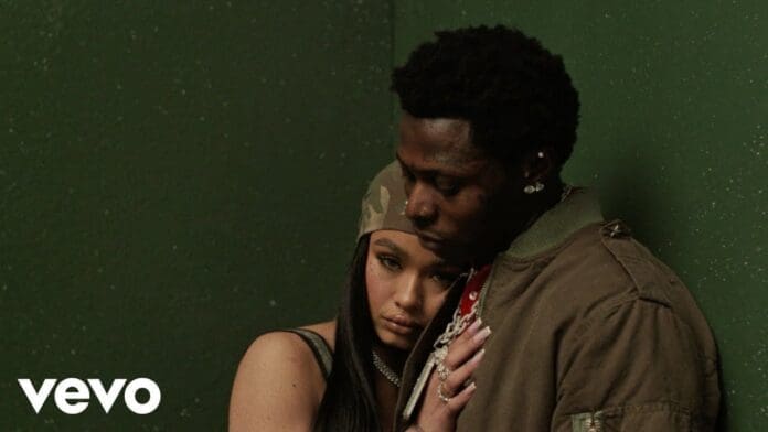 A woman rests her head on a man's chest as he embraces her; both have serious expressions and stand against a green wall. The Vevo logo appears in the lower left corner, marking this as the Official Video for Asake's "WHY LOVE.