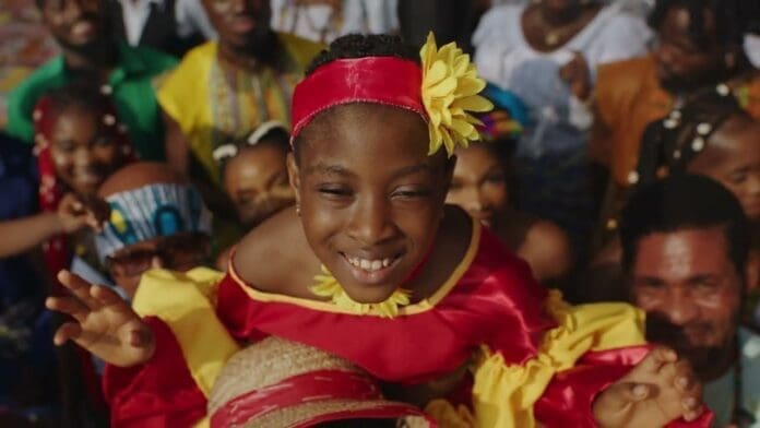 A smiling child in a red and yellow outfit with a yellow flower headband is lifted up by a crowd of people during a festive gathering, as Chiké's "Egwu" plays joyfully in the background.