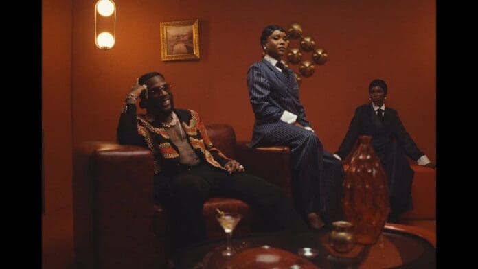 Three people in formal and stylish attire sit in a warmly lit, brown-toned room with modern and vintage decor—perfect for an Official Music Video update, complete with armchairs, art, and vases.