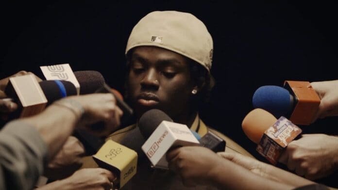 A young man wearing a beige cap looks down as multiple microphones are held toward him by reporters, suggesting a press conference or media interview, possibly related to Rema's Official Music Video for "DND.