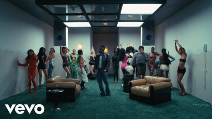 A man stands in the center of a modern room with green carpet, surrounded by people posing and dancing near the walls, moving to tunes by Wizkid; lounge chairs are in the foreground.