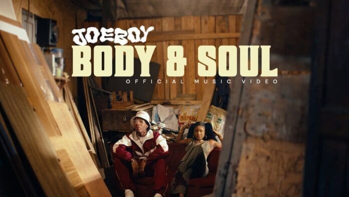 Two people sit in a dimly lit, cluttered wooden room as "Joeboy Body & Soul Official Video" text appears at the top of the image.
