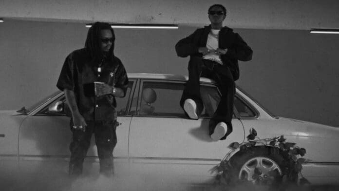 Two men pose by a parked car; one stands holding a drink, the other sits on the car roof with arms crossed. The black and white scene, with smoke and plants near the tire, channels the cool vibes of Skido and Olamide.