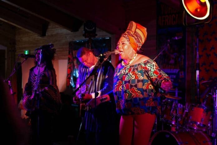 A band performs on stage under colorful lighting at the Yukon African Music Festival; the lead singer in vibrant patterned attire sings into a microphone while musicians play guitar and drums in the background.