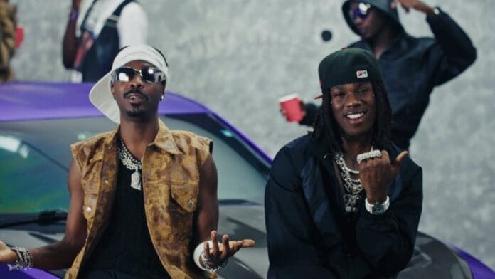 Rema and Young Jonn, both wearing stylish outfits and jewelry, pose in front of a purple car, with other people and a gray wall in the background.