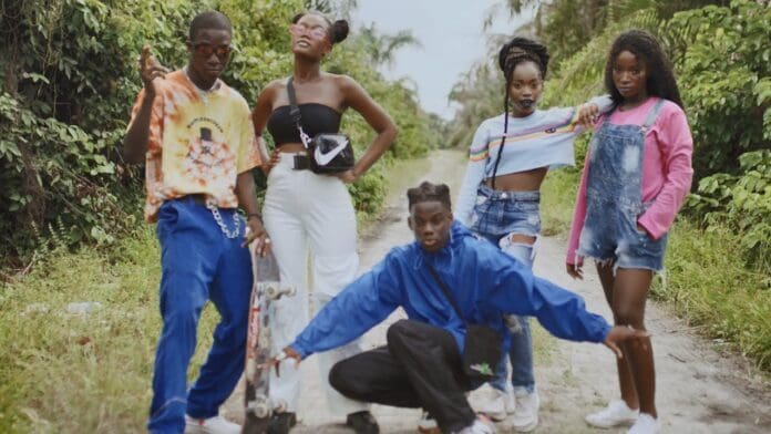 Five young people pose confidently on a dirt path surrounded by greenery, wearing colorful, casual outfits—one holds a skateboard and another squats in the foreground, capturing the vibrant energy of Rema's "Dumebi" Official Music Video.