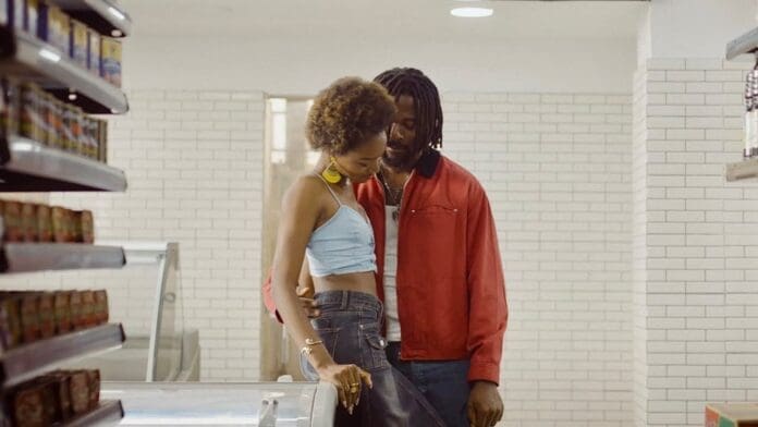 A man and woman stand closely together in a grocery store aisle, with the man’s arm around the woman’s waist; shelves and white tiled walls are visible, as Fireboy DML plays softly in the background.