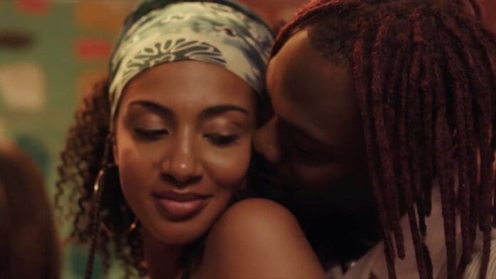 A woman with a headscarf smiles as a man with red hair leans in closely behind her in a warmly lit indoor setting, capturing the intimate vibe of CKay's "Emiliana" Official Music Video.