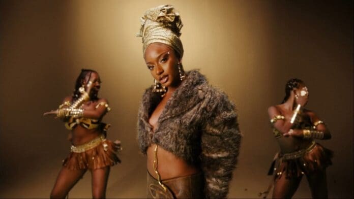 Ayra Starr shines in the Official Music Video for "Sability," wearing a fur jacket and gold headwrap as she stands before two dancers in brown and gold costumes, set against a gold-toned background.