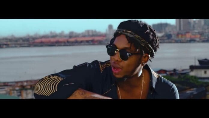 A man wearing sunglasses, a patterned headband, and a dark shirt sits outdoors with a city skyline and body of water in the background, reminiscent of the vibrant style seen in Runtown's "Mad Over You" Official Music Video.
