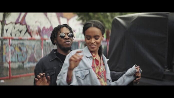 A man and woman dance together outdoors near a graffiti-covered wall and a fenced area in this relaxed and smiling scene from the Official Music Video for Runtown's "For Life." The man wears sunglasses as they enjoy the moment.