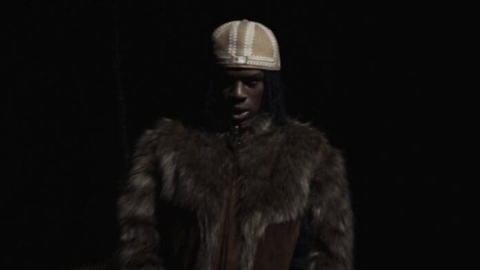 A person wearing a fur coat and a light-colored cap stands against a dark background, partially illuminated, as seen in the official music video for Rema’s track "OZEBA.