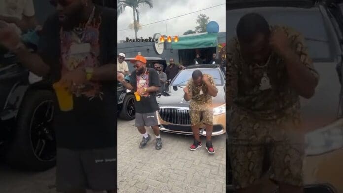 Two men dance LIVE in front of a luxury car as a crowd watches behind them outdoors; one holds a yellow cup, bringing DavidoOfficial vibes to the scene.