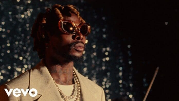 A man wearing large sunglasses, layered necklaces, and a beige suit stands in front of a shimmering, out-of-focus background in the Asake – “Remember” Official Video. The Vevo logo is in the bottom left corner.