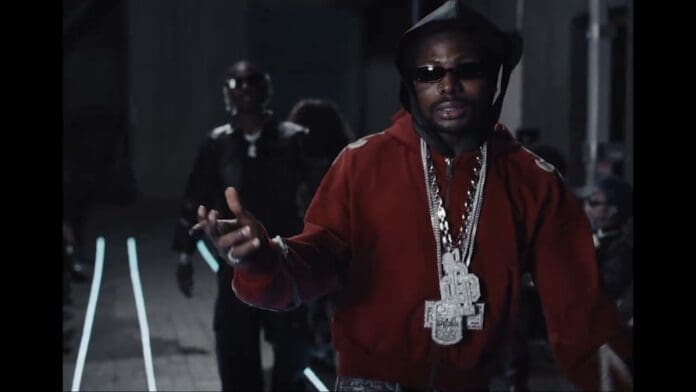 A man wearing a red hoodie, sunglasses, and multiple large chains gestures with his hand, channeling Young Jonn vibes, with another person blurred in the background in a dimly lit setting.