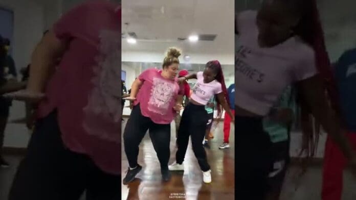 Two women in casual athletic wear take on the "Down Flat" Dance Challenge together in a mirrored studio, surrounded by other people, moving to the rhythms made popular by Kelvyn Boy.