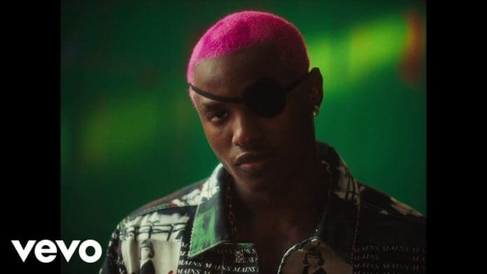 A person with bright pink hair and a black eye patch stands against a green background, wearing a patterned jacket inspired by Ruger’s bold style. Vevo logo is visible in the corner.