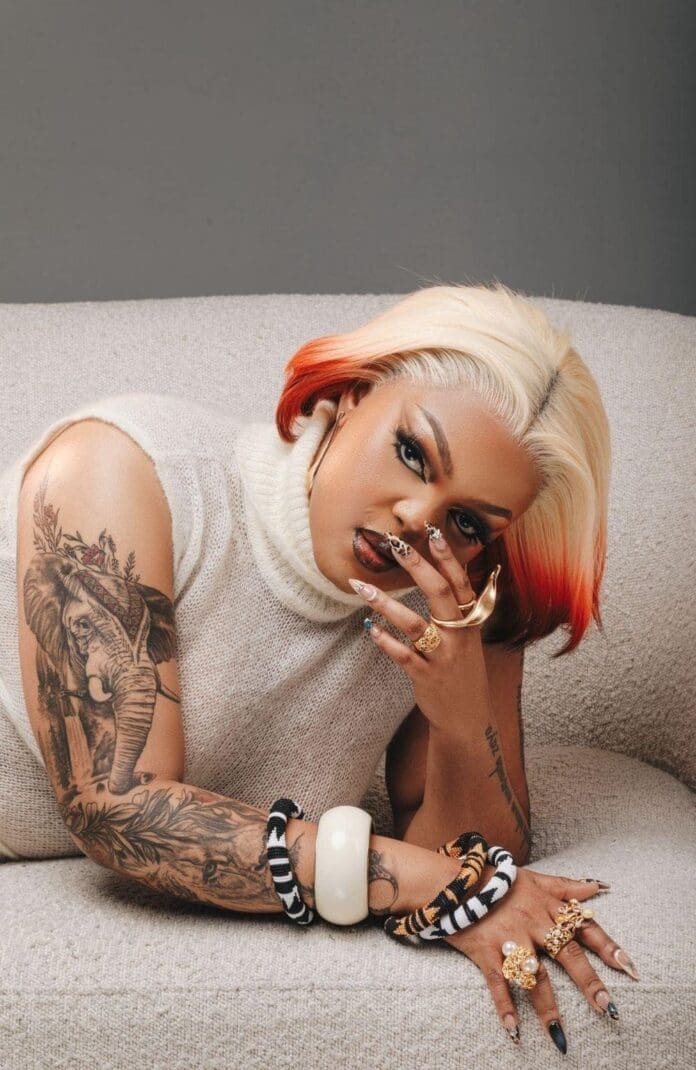 A female DJ with platinum blonde and red hair lies on a beige couch, showing tattoos, rings, and bracelets—one hand partially covering her face, reminiscent of a 24-hour set vibe.