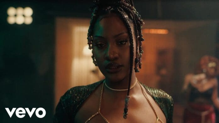 A woman with beaded braids and a shimmering outfit stands indoors, looking at the camera. Warm lighting and a blurred background set the mood as the Vevo logo and "Crazy Tings" by Tems indicate this is from the Official Video.