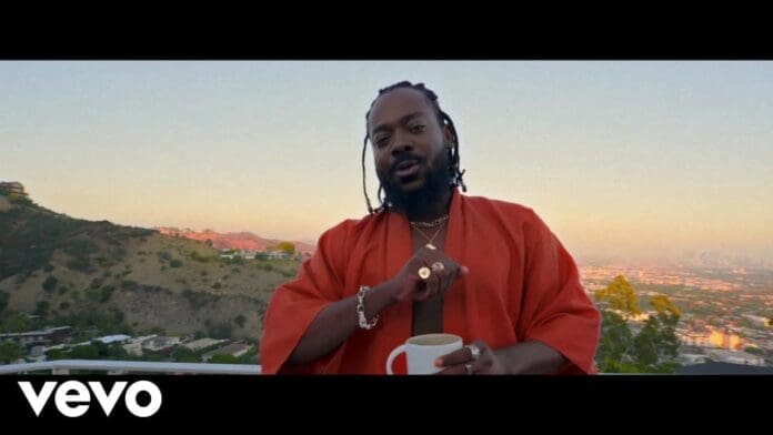 A man in a red robe holds a mug on a balcony overlooking a hilly cityscape at sunset. The Vevo logo appears in the corner, hinting at "Okay" by Adekunle Gold.