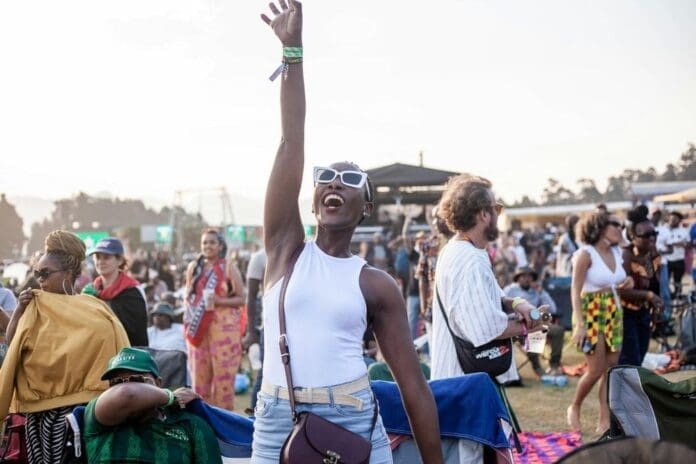 A solo female traveller in sunglasses and a white tank top raises her arm and smiles in a crowd at an outdoor Amapiano festival in South Africa, with people seated and standing around her.