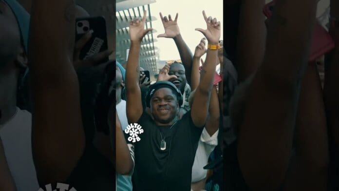 A group of people outdoors, with one person in the center wearing headphones and smiling, surrounded by others raising their hands in the air, enjoying an energetic amapiano music mix.