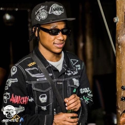 A person wearing a black Lakers cap, sunglasses, and a black jacket with various patches—possibly Rea Gopane—holds a drink and stands indoors, ready to join the global debate on trending issues.