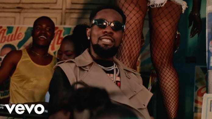 A man wearing sunglasses and a beige vest is in the foreground, with people and posters in the background; a person in fishnet stockings stands nearby. The Vevo logo marks this as the Official Video for "Abule" by Patoranking.