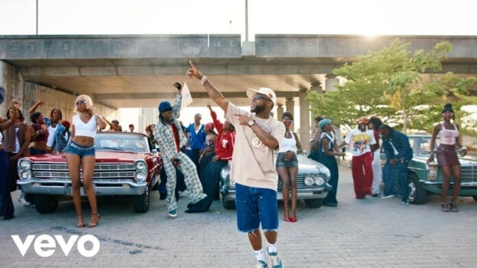 A group of people poses and dances in front of vintage cars under an overpass, with one man in the center pointing upward—capturing the vibrant energy of a Dance Video reminiscent of Davido’s “Be There Still.”.