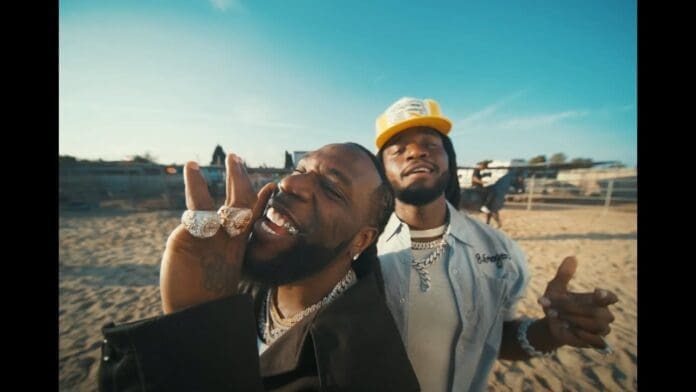 Two men, wearing jewelry and casual clothing, stand outdoors in a sandy, fenced area on a sunny day—smiling and gesturing towards the camera like they're ready to "Change Your Mind" with Shaboozey.