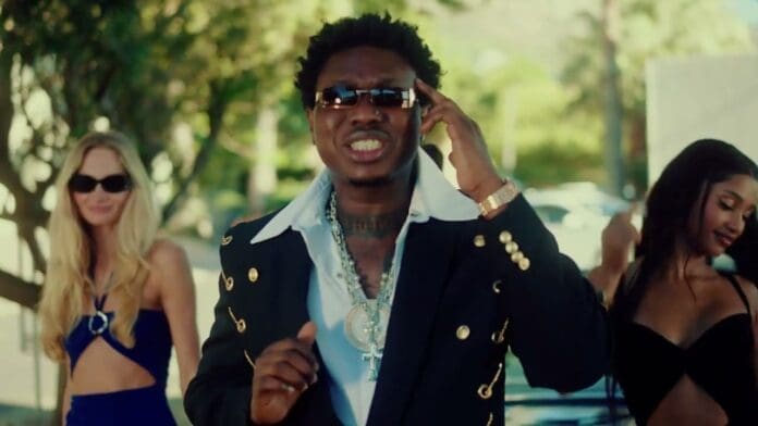 A man in a suit with gold buttons and sunglasses stands in the foreground, wearing jewelry, with two women in dresses and sunglasses behind him—capturing the glam vibe of a Zlatan "Happy Day" Official Video outdoors in daylight.