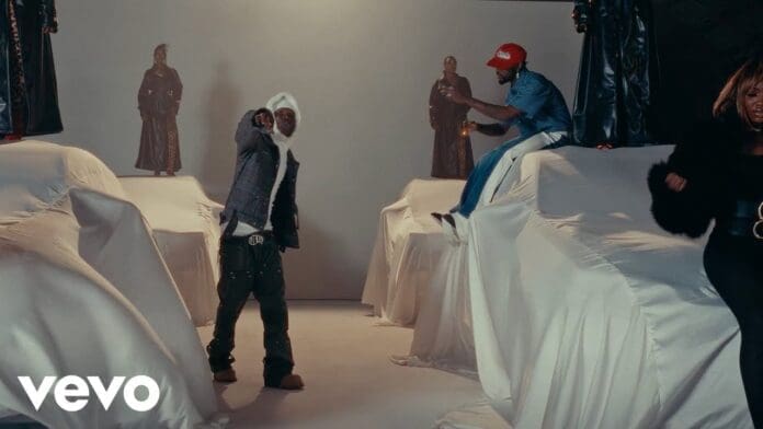 Two people pose on and beside large objects covered with white sheets in a studio setting, while a third person stands to the right. Statues are visible in the background, creating an atmosphere reminiscent of an Omah Lay "MY HEALER" music video.