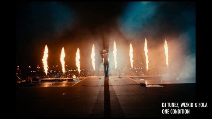 A performer stands on stage facing a large crowd, with tall flames shooting up behind them. Text in the corner reads: "DJ Tunez & Wizkid – One Condition.