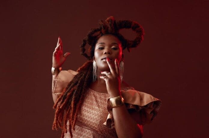 Nobuhle channels elegance as she poses with long textured hair and bold earrings against a plain brown background, wearing a patterned dress with ruffled sleeves and metallic jewelry—her style echoing the vibrant spirit of Amapiano.