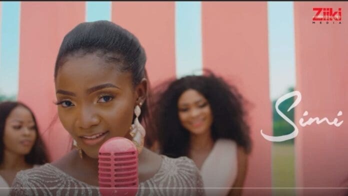A woman holding a pink microphone sings in front of two women, with pink panels and the name "Simi" in script on the right, capturing a moment from Simi's Official Video.