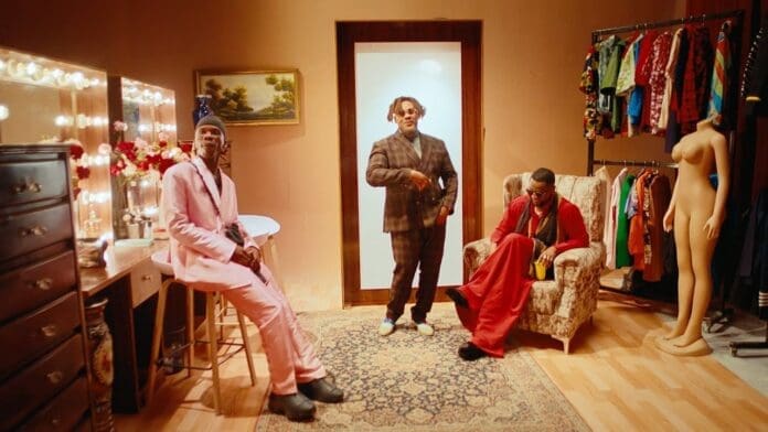 Three men pose in a stylish dressing room with a vanity, clothes rack, mannequins, and patterned rug on a wooden floor, channeling the vibrant vibe of GWAGWALADA by Kizz Daniel and BNXN fka Buju.