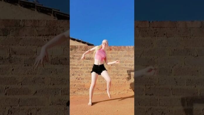 A person with long blond hair, wearing a pink crop top and black shorts, is barefoot and dancing the latest Amapiano Dance 2025 TikTok Challenge outside in front of a brick wall under a clear blue sky.