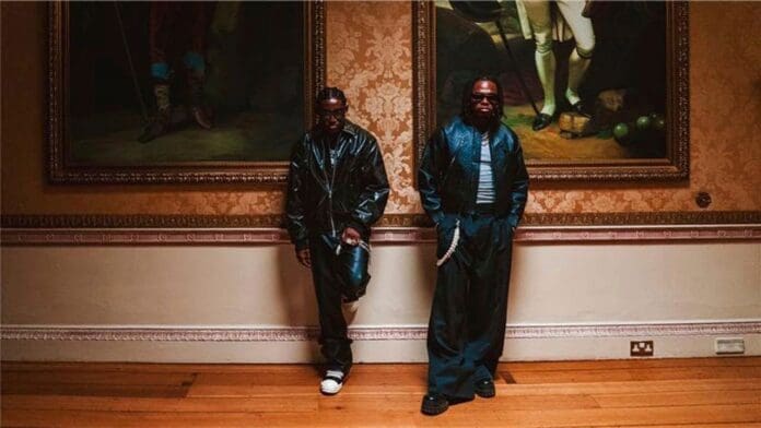 Two men, dressed in dark, shiny outfits reminiscent of Shallipopi and Gunna’s style, stand against an ornate wall adorned with framed paintings and wooden flooring in a formal room.