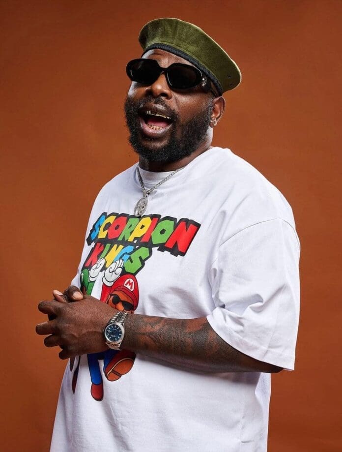 A man wearing sunglasses, a green beret, and a graphic t-shirt poses against a brown background with his hands clasped, evoking the spirit of unity often celebrated in South African hip hop.