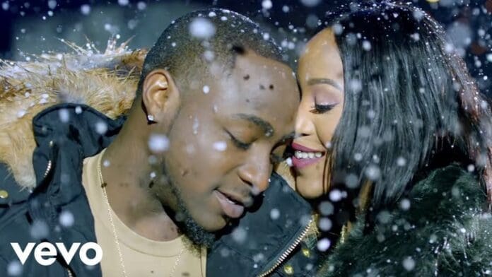 A man and a woman stand close together outside in falling snow, sharing an intimate moment. The scene feels like an Official Video for a heartfelt song—he closes his eyes as she smiles, leaning her face against his.