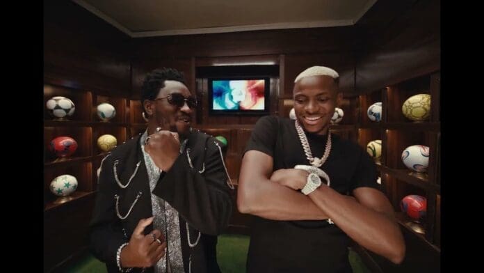 Two men stand smiling and posing in a room lined with shelves of soccer balls, filming the Friday Night Official Video, with a television screen in the background.