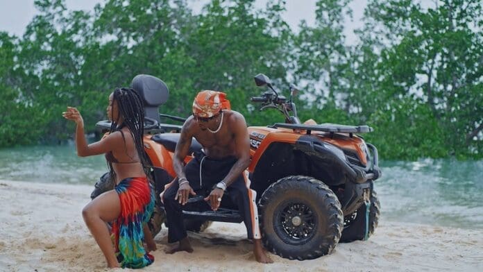 A woman and a shirtless man pose next to two orange ATVs on a sandy beach, with trees and water in the background, capturing the vibe of a Jeje Official Music Video.