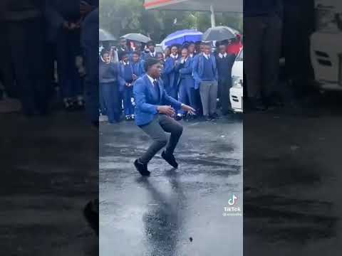 A student in a blue suit takes on a Dance Challenge, grooving energetically to Gqom beats on wet pavement as a crowd of people in uniforms and holding umbrellas watches.